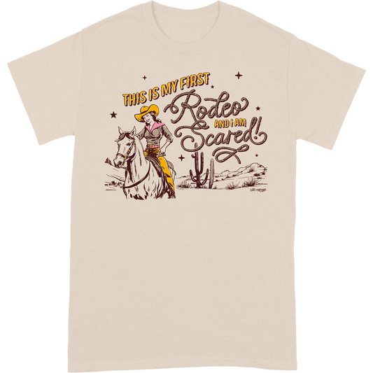 Light tan colored t-shirt that features artwork of a cowgirl riding a horse in the desert and the phrase "this is my first rodeo and I am scared"