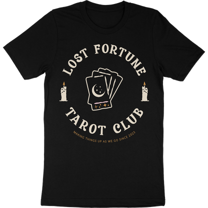 Black t-shirt with artwork of candles and a stack of tarot cards with a moon and stars on the front. The text reads "Lost Fortune Tarot Club. Making things up as we go since 2023." 