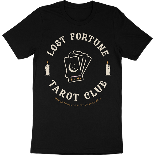 Black t-shirt with artwork of candles and a stack of tarot cards with a moon and stars on the front. The text reads "Lost Fortune Tarot Club. Making things up as we go since 2023." 