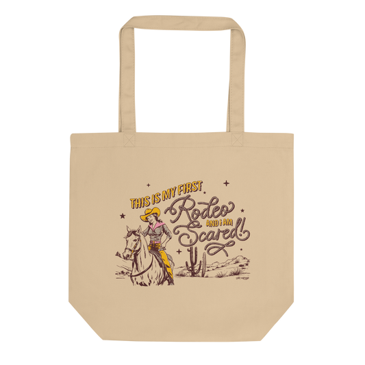 Tan tote bag that features artwork of a cowgirl riding a horse in the desert and the phrase "this is my first rodeo and I am scared"