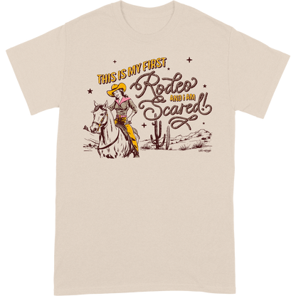 Light tan colored t-shirt that features artwork of a cowgirl riding a horse in the desert and the phrase "this is my first rodeo and I am scared"