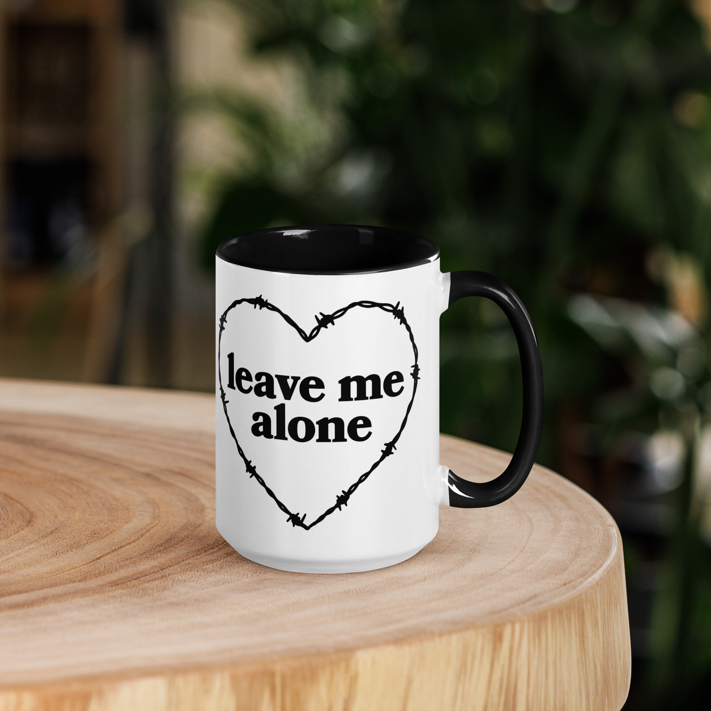 White ceramic mug with black handle and interior that features a barbed wire heart surrounding the phrase "leave me alone"